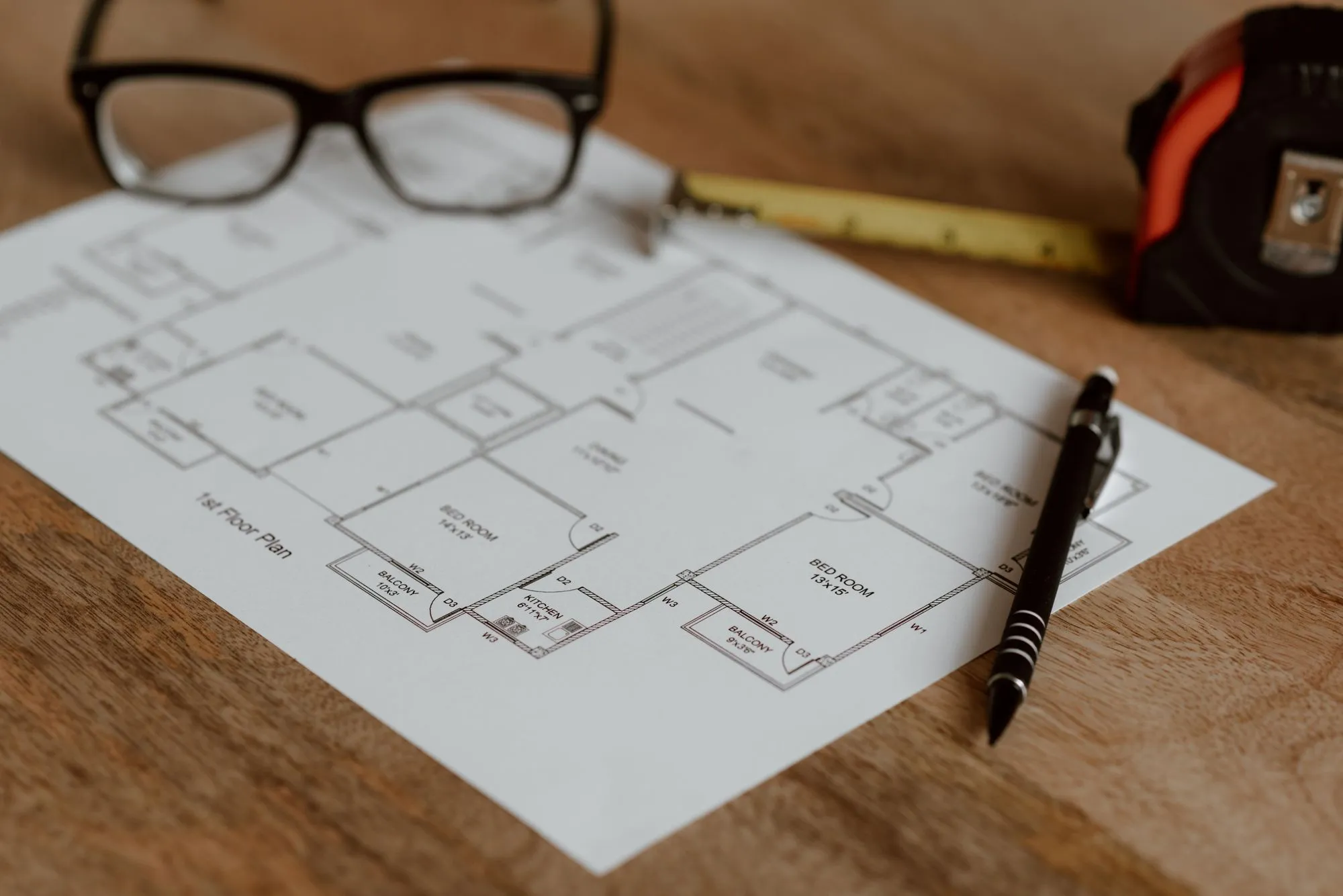 A house floor plan on a wooden table with a pen, glasses and a tape measure